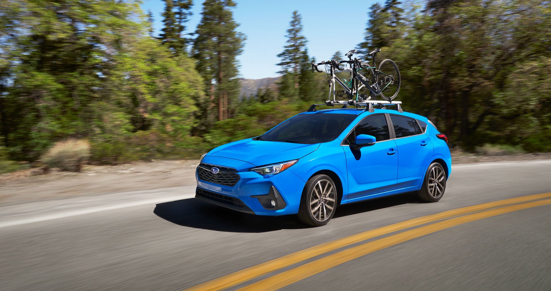 Blue 2024 Subaru hatchback driving in the mountains with bikes on roof rack.